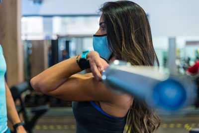 Side view of fit female in mask doing deadlift exercise with barbell under supervision of personal coach in modern gym