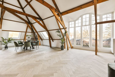 Round dining table with comfortable chairs in spacious light room with creative wood beams on ceiling and potted plants in modern apartment with terrace