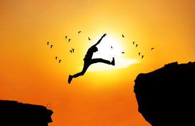 Full length of silhouette jumping on rock with birds flying against sky during sunset