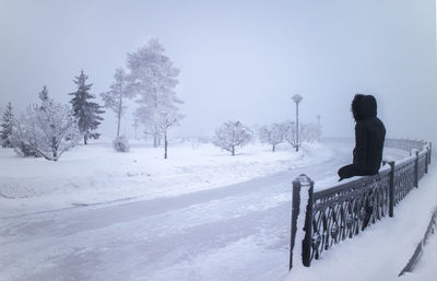 Hooded person sitting on railing by snow covered road against sky