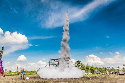 People looking at rocket while standing on field against blue sky