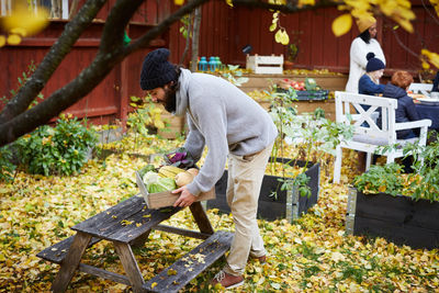 Man arranging vegetable basket on table while family and friends sitting in yard