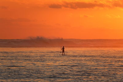 Silhouette man paddleboarding in sea against orange sky