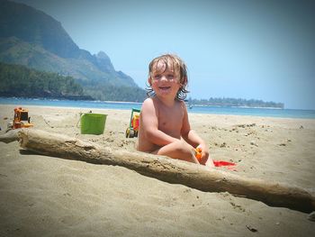 Happy shirtless boy sitting at beach against clear sky