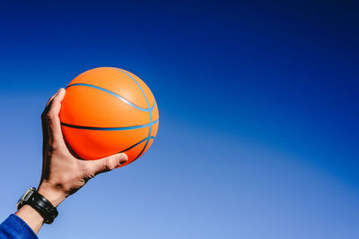 Cropped hand of man holding basketball against blue sky