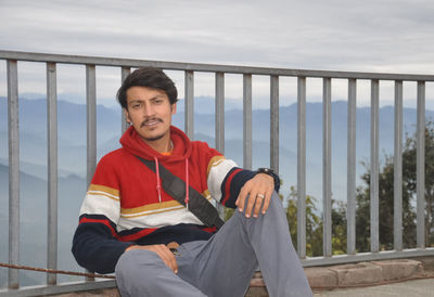 A handsome young guy looking at camera, sitting by safety barrier with the background of mountains