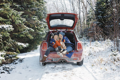 Cheerful female friends sitting in car trunk at algonquin provincial park during winter