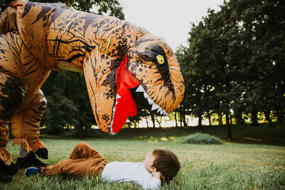 Boy lying on grassy land while playing with person wearing dinosaur costume in park