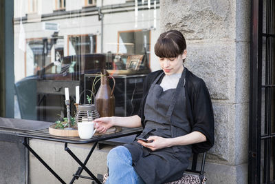 Young female owner using smart phone while sitting with coffee cup by table against store