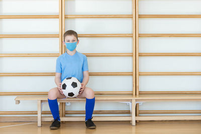 Portrait of boy wearing mask holding soccer ball sitting on bench