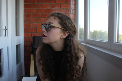 Portrait of young woman looking through window at home against brick wall