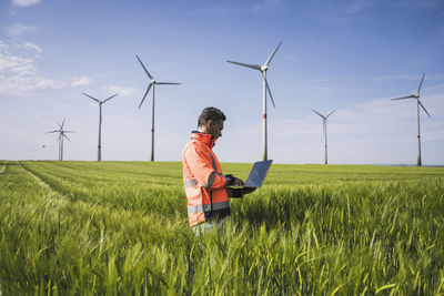 Engineer using laptop amidst wheat crops near wind turbines on field