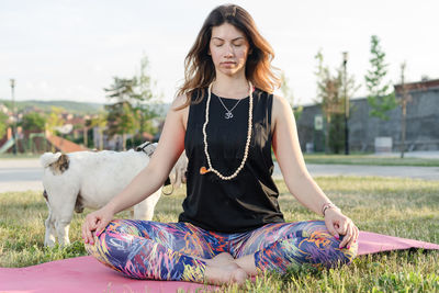 Woman meditating in lotus position while sitting by dog on grass