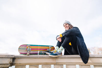 Low angle of full body of smiling young woman skater in casual outfit with skateboard sitting on border of stone fence