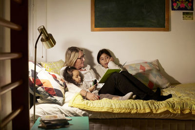 Grandmother reading book to granddaughters while sitting on bed at home