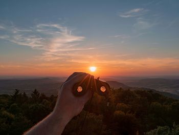 Cropped hand holding binoculars against sky during sunset