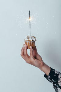 Cropped hand of woman holding cupcake with sparkler over white background