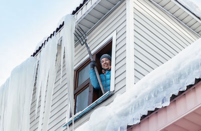 Smiling man removes giant icicles and snow from the roof using pitchfork through the window.