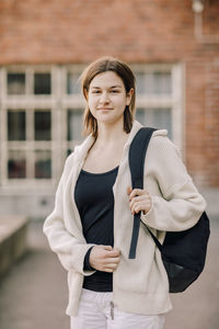 Portrait of smiling female teenage student with backpack standing in high school campus