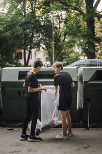 Full length of teenage boys holding garbage bag with plastic by recycling bin