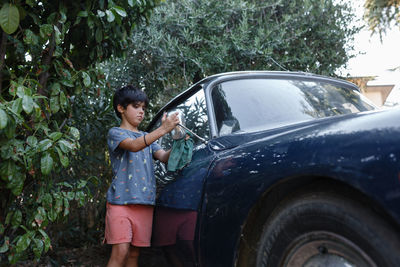 Girl of 8 years old with dark short hair washing the side mirror of blue vintage car