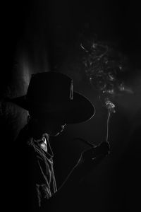 Obscured face of mature man holding cigarette while standing against black background