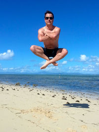 Full length of shirtless man jumping with crossed legged at beach