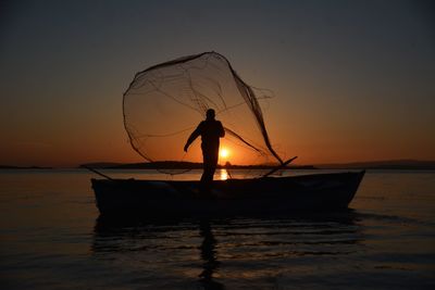 Silhouette man with fishing net standing in boat on sea during sunset