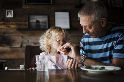 Grandfather feeding food to granddaughter while sitting at table in home