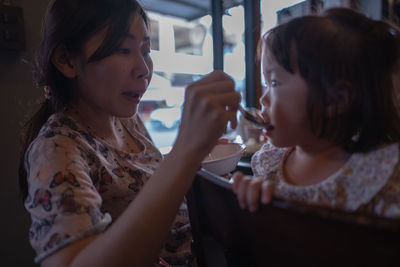 Woman feeding toddler daughter sitting on chair in restaurant