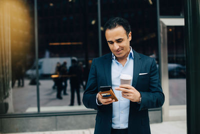 Smiling businessman using smart phone while holding disposable coffee cup against building in city