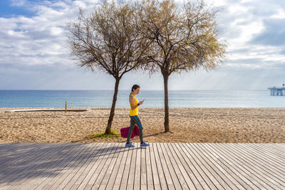 Side view of sportive woman walking on promenade while using mobile