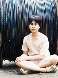 Portrait of young man sitting on footpath against rolled cable wires