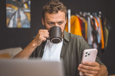 A man looking at his cell phone while holding a coffee mug.