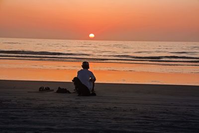 Man sitting on beach against orange sky