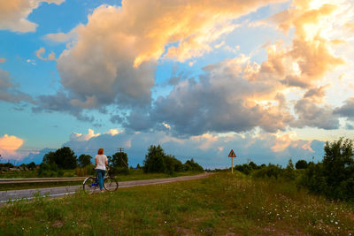 Rear view of woman with bicycle amidst field on road against sky