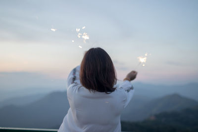 Rear view of woman holding sparklers against sky during sunset