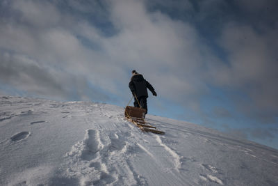 Low angle view of man pulling sled while walking on snow covered mountain against cloudy sky