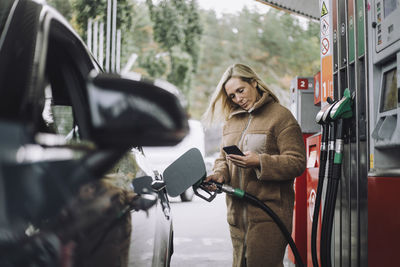 Woman using mobile phone while refueling car at gas station