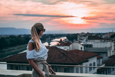 Woman looking at cityscape against sky during sunset