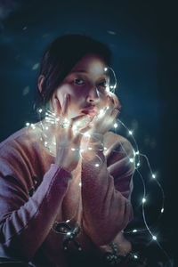Portrait of beautiful young woman holding illuminated string lights while sitting against wall