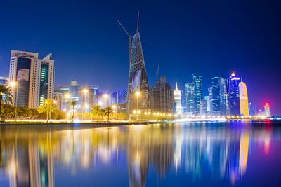 Illuminated buildings by river against clear blue sky at night