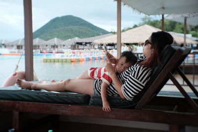 Woman with son lying on lounge chair while using mobile phone at beach