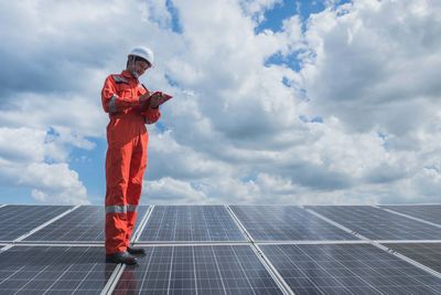 Low angle view of worker wearing reflective clothing while standing on solar panel against sky