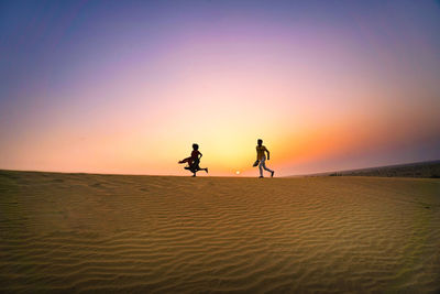 Silhouette people riding on desert against sky during sunset