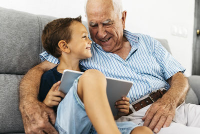 Happy little boy with digital tablet sitting beside his grandfather on the couch at home having fun
