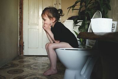 Side view of girl sitting on toilet bowl in bathroom at home