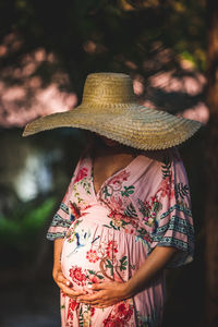 Pregnant woman, in pink dress, side view of woman wearing hat standing outdoors.