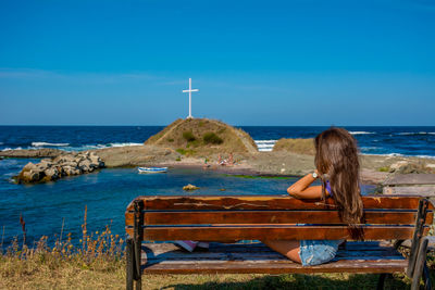 Side view of mid adult woman sitting on bench at beach against clear blue sky during sunny day