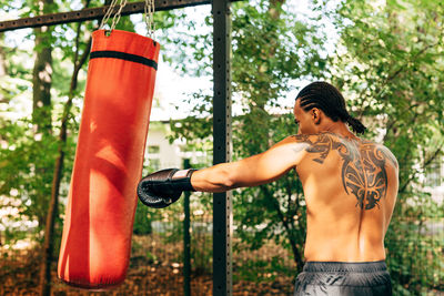 Rear view of shirtless man boxing while standing against tree